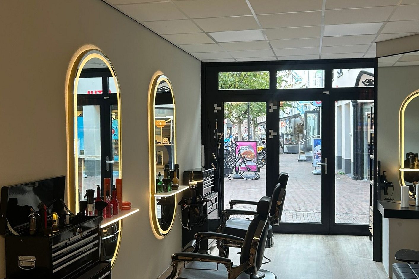 Het interieur van Schaar & Ziel: verlichte ovale spiegels, kappersstoelen en de glazen deur naar de Markt